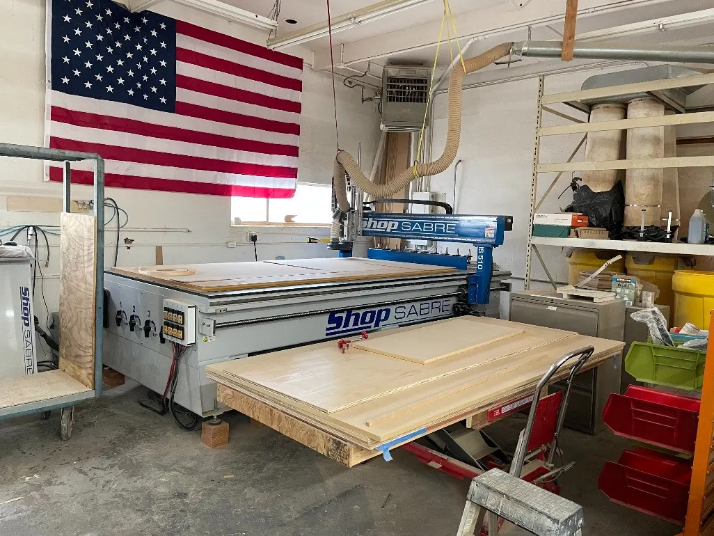 The 5x10 CNC router cutting a sheet in the NR Woodworks shop in Camarillo, an American flag on the wall