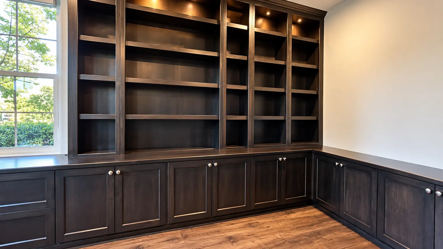 Dark stained built in library with lit shelves above paneled cabinets