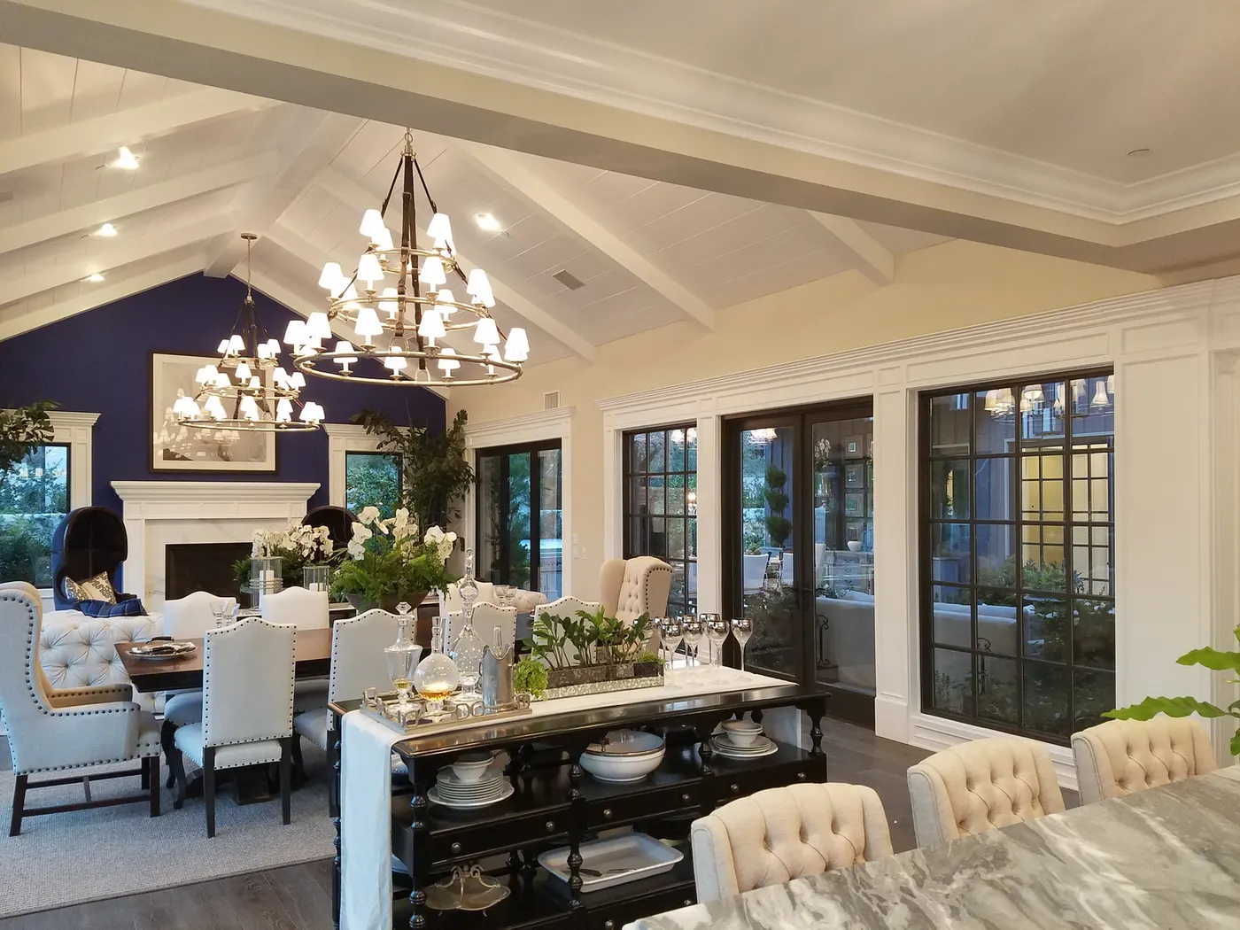 Great room with a navy custom kitchen, a dining table under a chandelier and a white fireplace wall